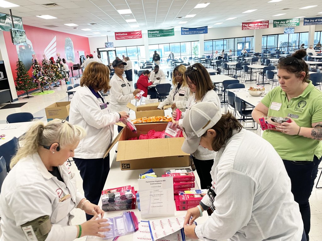 A group of people packing items into boxes, with tables and holiday decorations in the background.