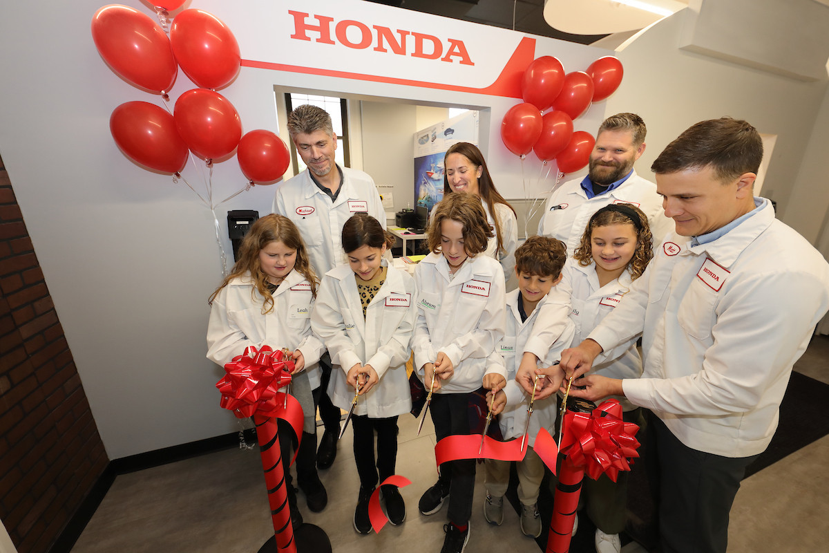 a group of students and Honda employees wearing white coats, cutting a red ribbon together with red balloons in the background
